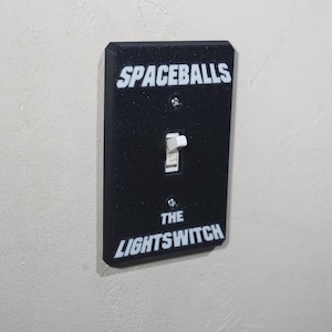 May include: Black light switch cover with the text "SPACEBALLS" above the switch and "THE LIGHTSWITCH" below it in white lettering. The switch is in the on position.
