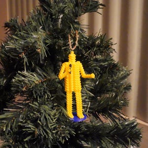 May include: A yellow, spiral-shaped pasta figure Christmas tree ornament. The figure has a blue base and a brown string for hanging. The ornament is hanging on a green Christmas tree.