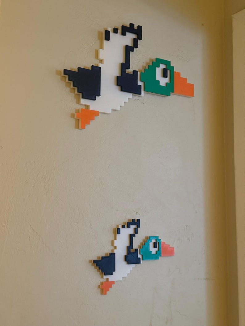 May include: Two pixel art duck figures on a beige wall. The larger duck is white with black wings, a green head, and an orange beak. The smaller duck is the same design but smaller in size.