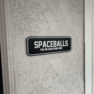 May include: A rectangular black sign with a white border featuring the white text SPACEBALLS in a bold font and the smaller white text THE NO SOLICITING SIGN underneath. The sign is mounted on a textured off-white wall surface.