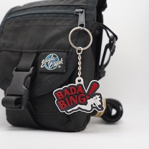 Può includere: Un marsupio nero con cerniera e fibbia argentate. Un portachiavi con le parole "Bada Bing" in rosso e bianco, e un design di mazza da baseball, pende dalla cerniera.