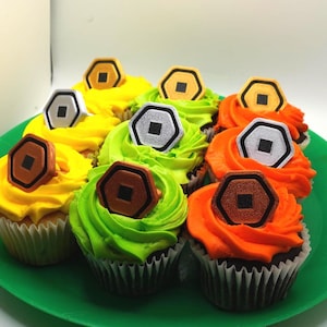 May include: A collection of cupcakes with colorful frosting in yellow, green, and orange. Each cupcake is topped with a decorative hexagon-shaped emblem in various colors. The cupcakes are arranged on a green plate.