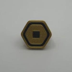 May include: A hexagonal, gold-colored button with a black border and a black square in the center. The button is set against a white background. The button's design is geometric and symmetrical.