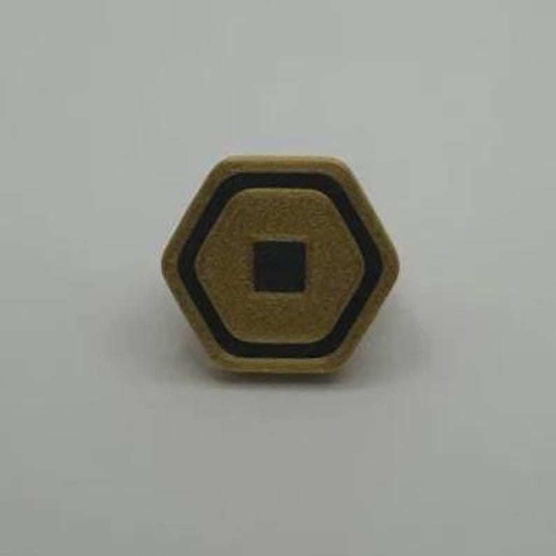 May include: A hexagonal, gold-colored button with a black border and a black square in the center. The button is set against a white background. The button's design is geometric and symmetrical.