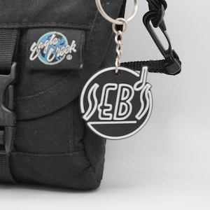 May include: Black bag with a silver keychain featuring a black and white circular charm. The charm has the text "SEB's" in a stylized font, with a musical note. The bag has the "Eagle Creek" logo.