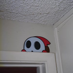 Pode incluir: Um adesivo de desenho animado de um personagem Shy Guy espreitando por cima de uma moldura de porta branca. O personagem tem uma máscara cinza com olhos grandes e pretos, um capuz vermelho e um corpo marrom. O adesivo é colocado em uma parede branca.