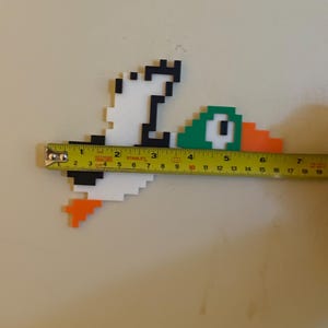 May include: A pixel art duck made of white, black, green, and orange plastic blocks. The duck is approximately 7 inches long.