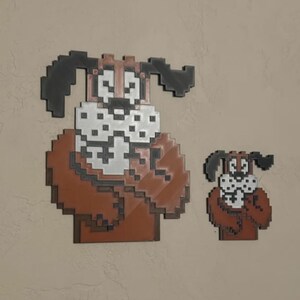 May include: Two pixel art dog wall decorations. The larger dog has a brown body, white face, and black ears. The smaller dog has the same design. Both are mounted on a neutral-coloured wall.