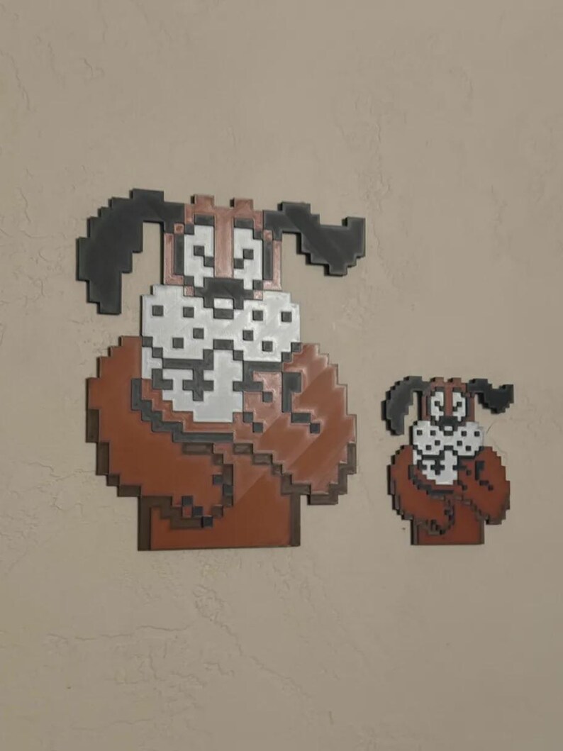 May include: Two pixel art dog wall decorations. The larger dog has a brown body, white face, and black ears. The smaller dog has the same design. Both are mounted on a neutral-coloured wall.