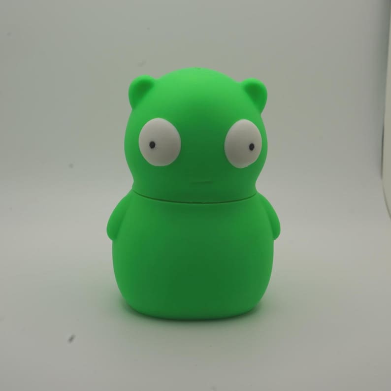 May include: A bright green, cartoon-like figure with large white eyes and small black pupils. The figure has a rounded body, small arms, and two small ears. The object is a solid, smooth, and rounded shape, likely a container or a toy.