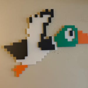 May include: A pixelated duck with a green head, orange beak, and black and white wings is flying. The duck is made of wood and is mounted on a wall.