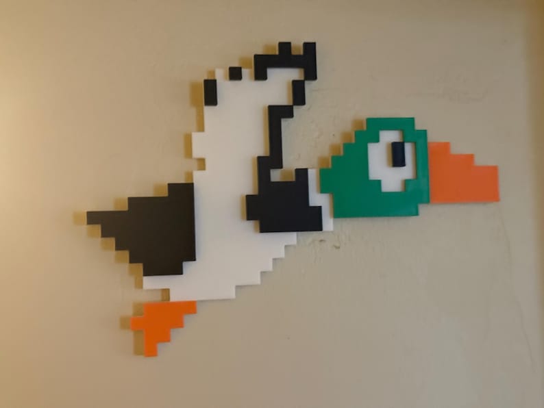 May include: A pixelated duck with a green head, orange beak, and black and white wings is flying. The duck is made of wood and is mounted on a wall.