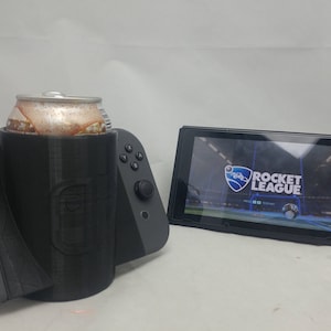 May include: A black and gray Nintendo Switch console with a gray Joy-Con controller on the left and a black Joy-Con controller on the right. A black can holder is attached to the console, holding a can of soda. The console is displaying a video game screen with the text "ROCKET LEAGUE".