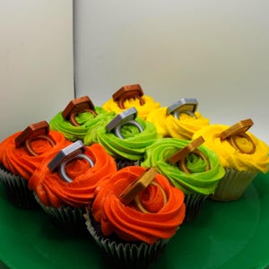 May include: A collection of cupcakes with colorful frosting, including orange, green, and yellow. Each cupcake is topped with a decorative ring, creating a festive and celebratory appearance. The cupcakes are arranged on a green plate.
