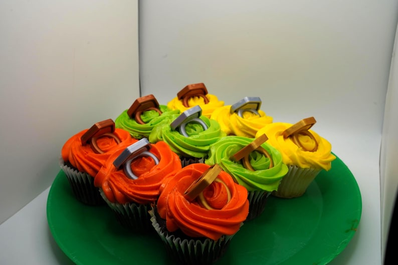 May include: A collection of cupcakes with colorful frosting, including orange, green, and yellow. Each cupcake is topped with a decorative ring, creating a festive and celebratory appearance. The cupcakes are arranged on a green plate.
