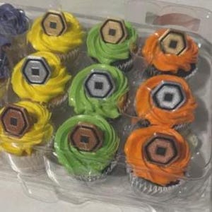 May include: A clear plastic container holds a dozen cupcakes with colorful frosting. The frosting colors are yellow, green, and orange. Each cupcake is topped with a brown and silver hexagonal decoration. The cupcakes are arranged in rows of three.