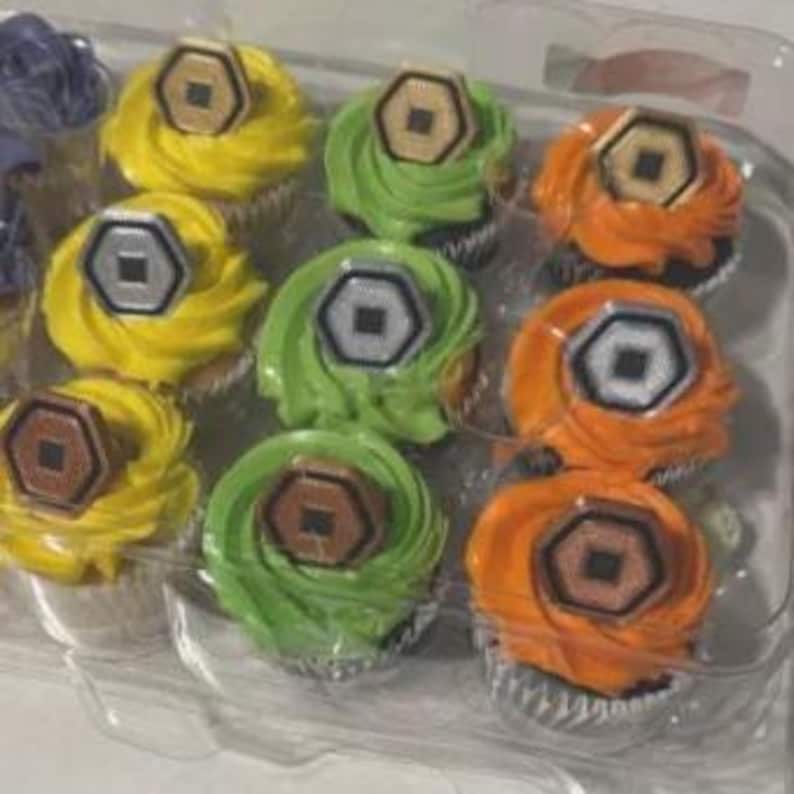 May include: A clear plastic container holds a dozen cupcakes with colorful frosting. The frosting colors are yellow, green, and orange. Each cupcake is topped with a brown and silver hexagonal decoration. The cupcakes are arranged in rows of three.