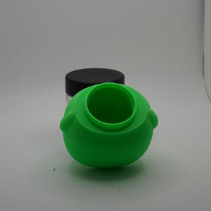 May include: A bright green silicone container with a round body and a wide opening. The container has two small, rounded protrusions on either side. A clear jar with a black lid is in the background.