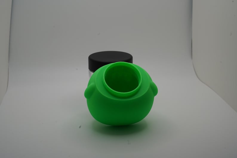May include: A bright green silicone container with a round body and a wide opening. The container has two small, rounded protrusions on either side. A clear jar with a black lid is in the background.