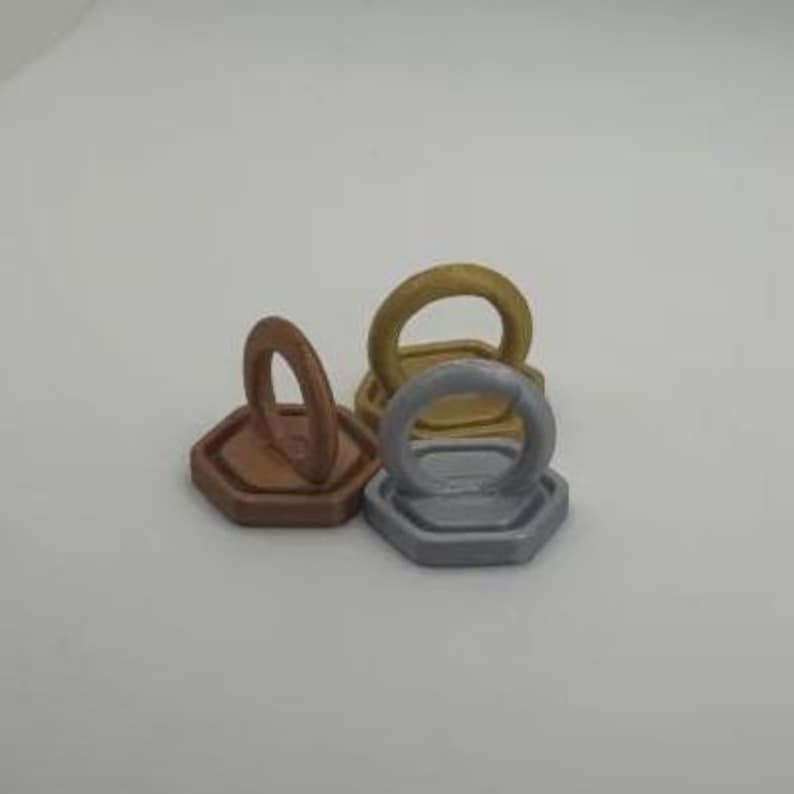 May include: A collection of four metallic phone grips in bronze, gold, and silver. Each grip features a hexagonal base with a circular ring attached, designed to enhance phone handling and provide a secure grip.