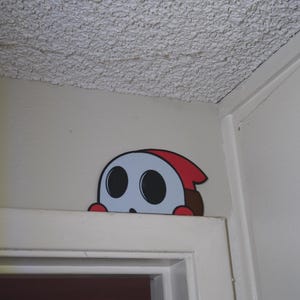 May include: A flat, decorative wall decal featuring a cartoon character with a pale blue mask, large black oval eyes, a small black mouth, and a red hood. The character is positioned to appear as if it is peeking over the top of a door frame.