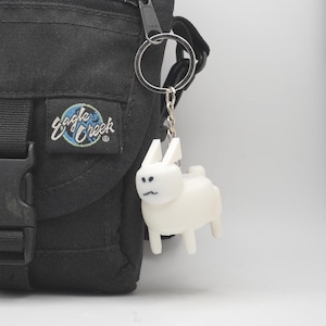 Könnte beinhalten: Ein weißer Hasen-Schlüsselanhänger mit einem silbernen Schlüsselring und einer Kette hängt an einer schwarzen Tasche. Der Hase hat ein minimalistisches Design mit kleinen Ohren und einem einfachen Gesicht. Die Tasche hat einen "Eagle Creek"-Aufnäher.