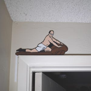 May include: A decorative wooden cutout of a man in underwear and glasses, reclining on a brown couch. The cutout is mounted on a white door frame, with the man's image extending over the top of the door.