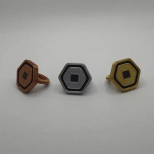 May include: Three rings with a geometric design. The rings are in bronze, silver, and gold colors. Each ring has a hexagonal face with a square center and a black outline. The bronze ring has a band, while the others are flat.