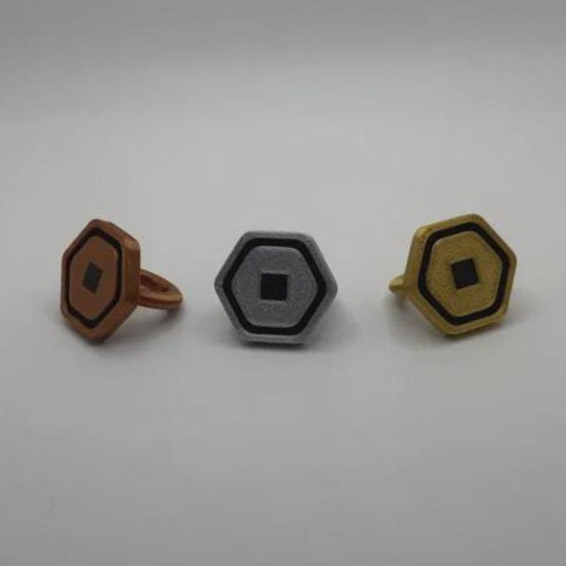 May include: Three rings with a geometric design. The rings are in bronze, silver, and gold colors. Each ring has a hexagonal face with a square center and a black outline. The bronze ring has a band, while the others are flat.