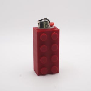 May include: A red lighter encased in a red building block design. The lighter has a silver metal top with a red ignition button. The building block design features eight red circular studs.