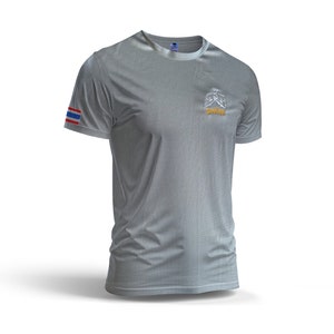 Puede incluir: Una camiseta gris de manga corta con un sutil patr&oacute;n texturizado. La camiseta presenta una peque&ntilde;a bandera tailandesa en la manga izquierda y un logotipo con texto en el pecho. La camiseta se muestra sobre un fondo blanco.