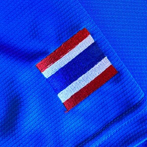 Puede incluir: Primer plano de una tela azul vibrante con un patr&oacute;n texturizado. Un parche rectangular con la bandera tailandesa, con rayas rojas, blancas y azules, est&aacute; cosido a la tela. La bandera est&aacute; bordada con puntadas detalladas.