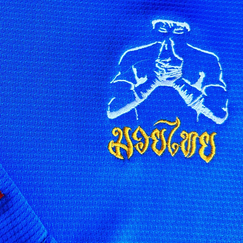 Puede incluir: Primer plano de una camiseta deportiva azul con un contorno bordado blanco de una persona en posici&oacute;n de oraci&oacute;n. Debajo de la figura hay texto dorado en una fuente estilizada. La camiseta tiene una superficie texturizada.