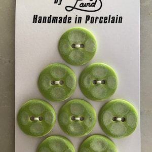 May include: A set of eight green porcelain buttons with white accents. The buttons are arranged in a triangle shape on a white card with the text "DESIGNER BUTTONS by Laud Handmade in Porcelain".