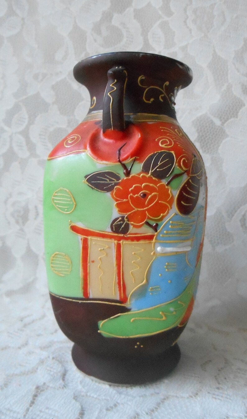 May include: A small, hand-painted ceramic vase with a green, red, blue, and gold floral design. The vase has a brown base and a handle on the side.