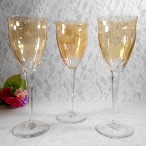 May include: Three elegant, clear wine glasses with a golden-amber hue. The glasses have a tall, slender stem and a wide, flared bowl. The base is round and clear. The glasses are arranged on a white lace surface.
