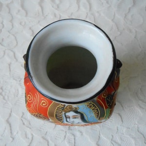 May include: A small, red and gold ceramic vase with a white interior and a black rim. The vase is decorated with a Japanese-inspired design featuring a woman's face and floral patterns.