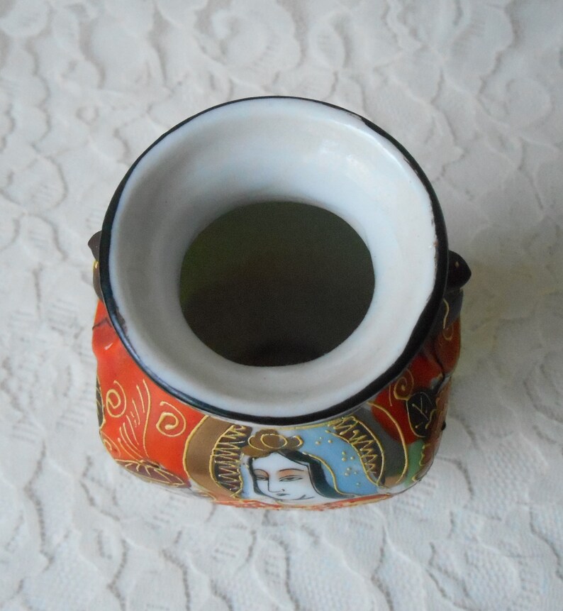 May include: A small, red and gold ceramic vase with a white interior and a black rim. The vase is decorated with a Japanese-inspired design featuring a woman's face and floral patterns.