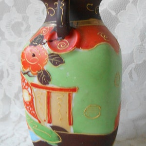 May include: A small, brown and green ceramic vase with a floral design and gold accents. The vase has a handle on one side and a rounded base.