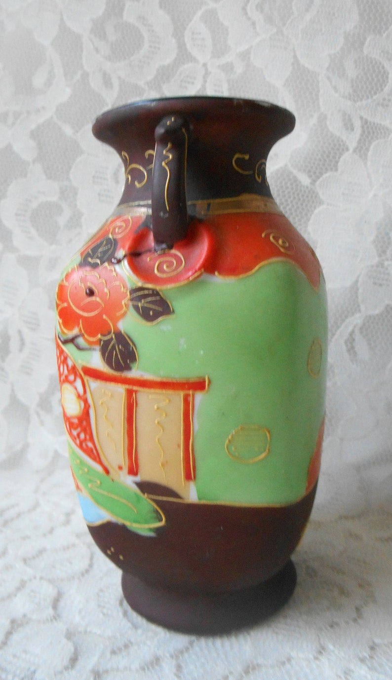 May include: A small, brown and green ceramic vase with a floral design and gold accents. The vase has a handle on one side and a rounded base.