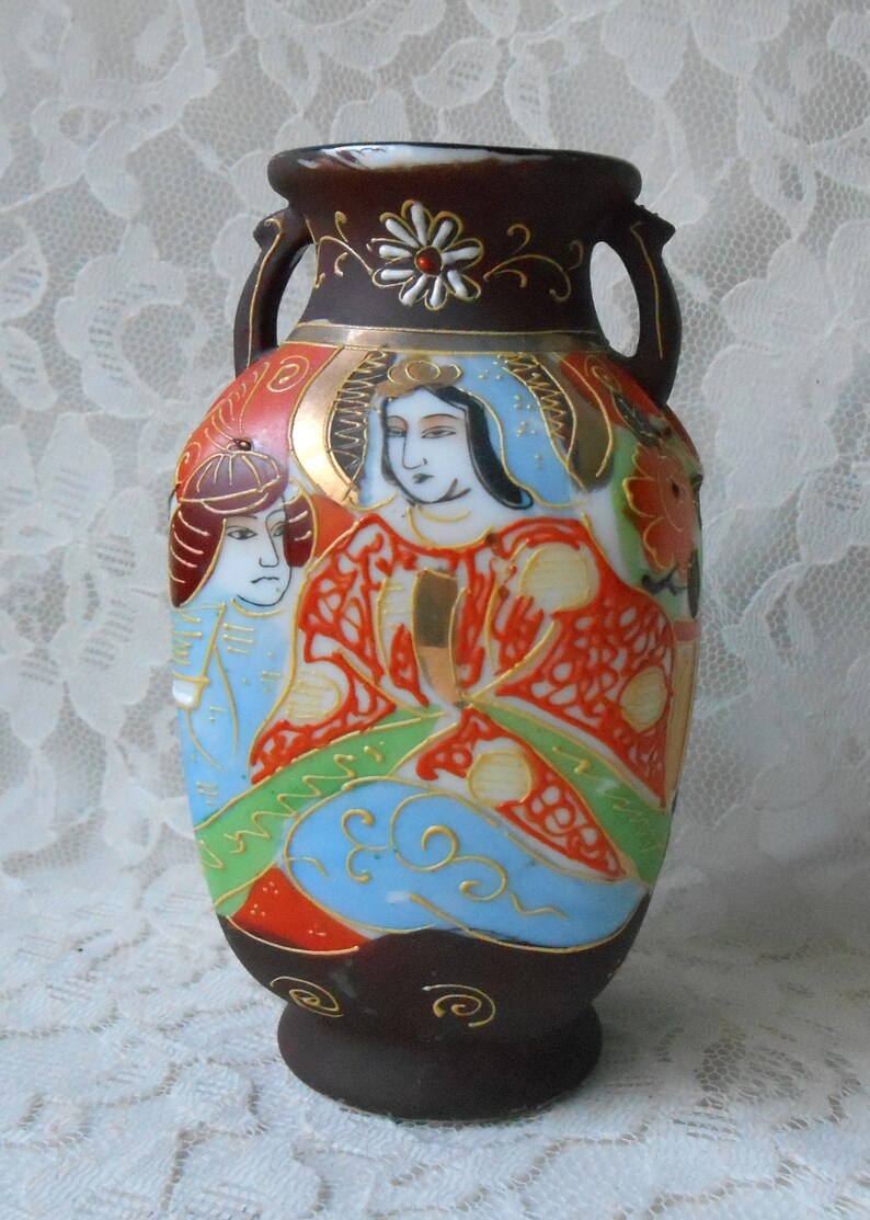 May include: A small, dark brown ceramic vase with two handles. The vase is decorated with a colorful Japanese-inspired design featuring three figures in traditional clothing. The figures are painted in shades of red, blue, green, and gold.