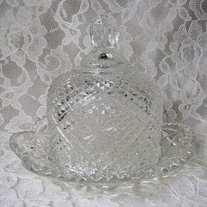 May include: A clear, cut-glass butter dish with a domed lid and a matching round base. The dish features a diamond-cut pattern and a decorative finial on top. The base has a scalloped edge. A vintage kitchenware item.