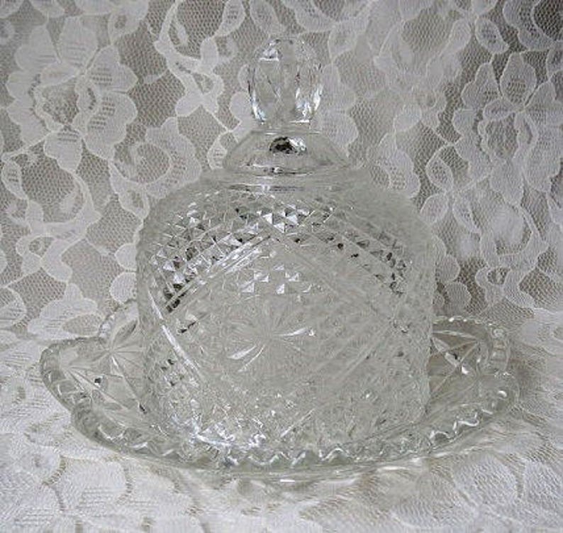 Pode incluir: Uma manteigueira de vidro transparente lapidado com tampa abobadada e base redonda a condizer. O prato apresenta um padr&atilde;o de corte de diamante e um adorno decorativo no topo. A base tem uma borda ondulada. Um artigo de cozinha vintage.