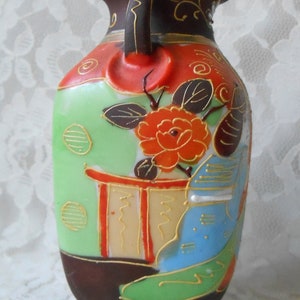 May include: A small, hand-painted ceramic vase with a green, red, blue, and gold floral design. The vase has a brown base and a handle on the side.