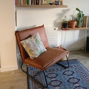 Puede incluir: Un sill&oacute;n de cuero marr&oacute;n con un marco de metal negro se encuentra sobre una alfombra de patr&oacute;n azul y blanco. Un coj&iacute;n blanco con estampado floral verde est&aacute; en el sill&oacute;n.