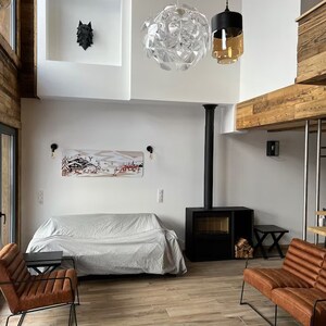 May include: A living room with a white couch, a black wood-burning stove, and two brown leather armchairs. The room has a high ceiling with a large, clear glass chandelier and a black wolf sculpture on the wall.