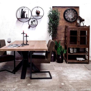 May include: A wooden dining table with a black metal base and two chairs. The table has a natural wood finish and the chairs have brown upholstery. There are three black candle holders on the table. The table is set against a white wall with three round black metal shelves. The shelves are decorated with books and a globe. There is a wooden cabinet with glass doors and shelves on the right side of the image. The cabinet is decorated with a clock and a lamp.