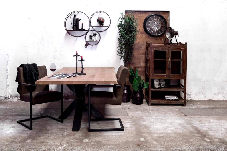 May include: A wooden dining table with a black metal base and two chairs. The table has a natural wood finish and the chairs have brown upholstery. There are three black candle holders on the table. The table is set against a white wall with three round black metal shelves. The shelves are decorated with books and a globe. There is a wooden cabinet with glass doors and shelves on the right side of the image. The cabinet is decorated with a clock and a lamp.