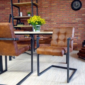 May include: Four brown leather chairs with black metal frames and a wooden table in a room with a brick wall. The chairs have a tufted design and are arranged around the table.