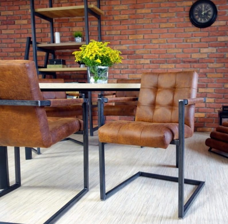 May include: Four brown leather chairs with black metal frames and a wooden table in a room with a brick wall. The chairs have a tufted design and are arranged around the table.
